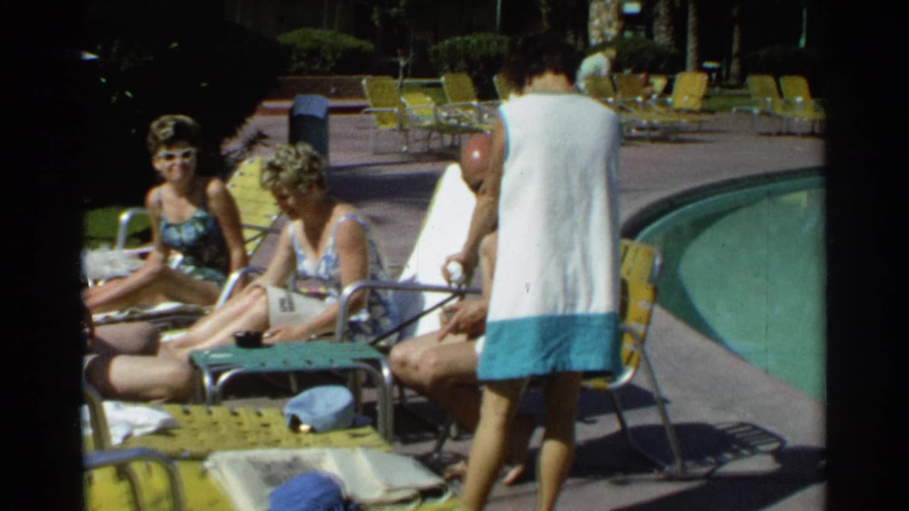 Adults in swimwear sit on yellow lounge chairs beside a pool while a woman in a cover-up walks past.