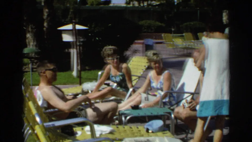 Scottsdale 1968 home-movie frame of adults lounging and talking near the pool, with shaded landscaping and yellow chairs around them.