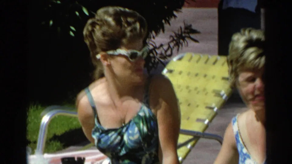 Close poolside view of two adult women in patterned 1960s swimsuits, one wearing white sunglasses, seated near yellow lounge furniture.