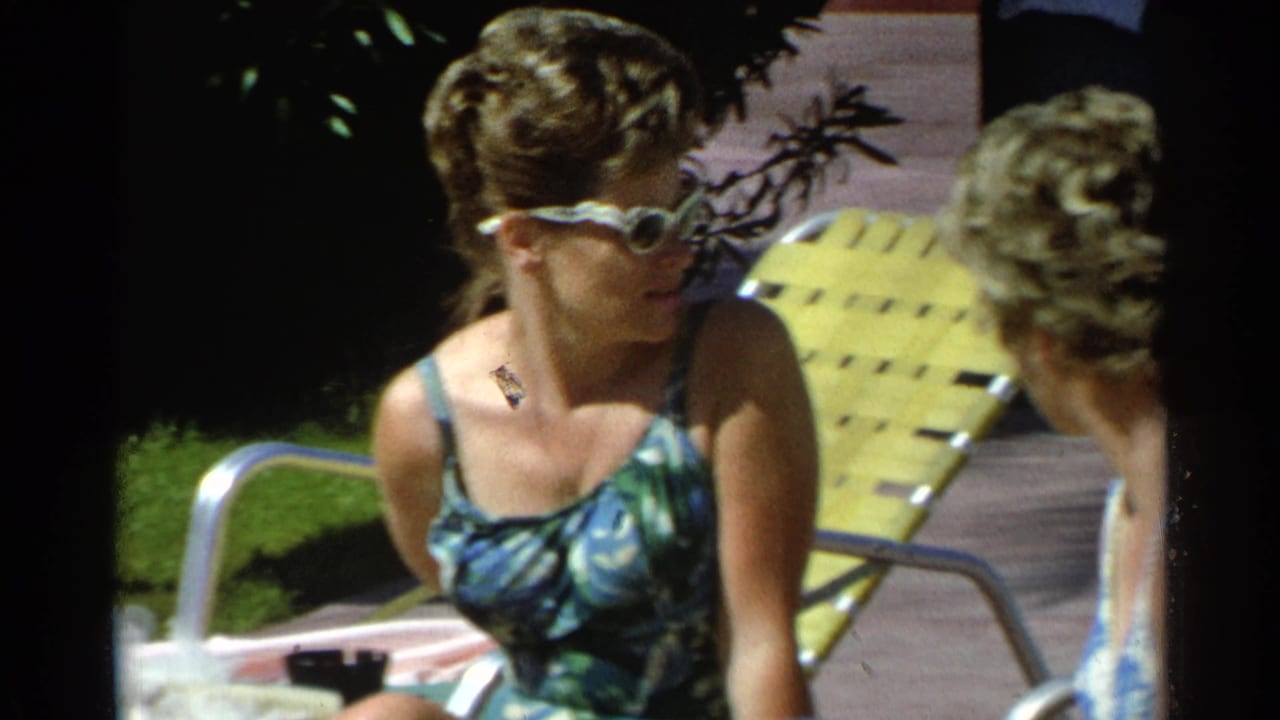 Woman in sunglasses sits near a poolside lounge chair beside another adult.