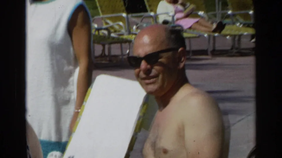 A shirtless man wearing dark sunglasses smiles at the Scottsdale poolside, with lounge chairs and another adult nearby.
