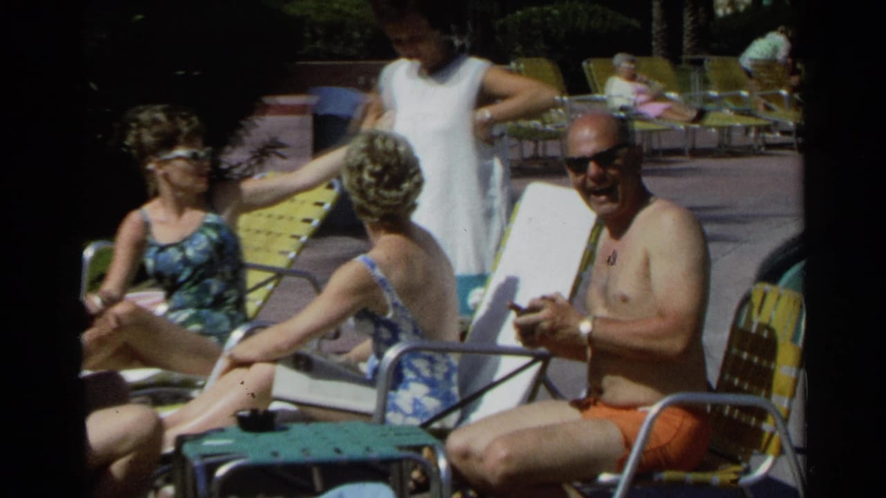 Adults in swimsuits sit on metal lounge chairs beside a sunny pool area.