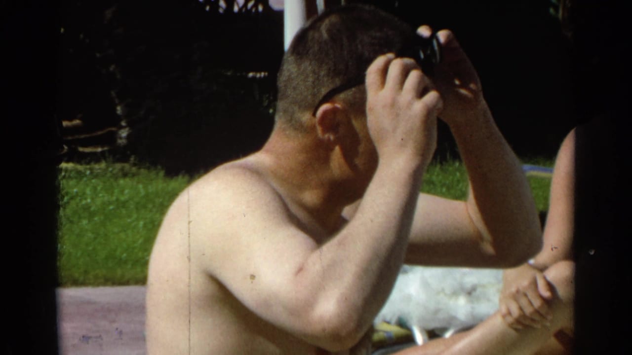 In 1968 Scottsdale home-movie footage, a shirtless man adjusts sunglasses while seated near grass beside a partially visible person.