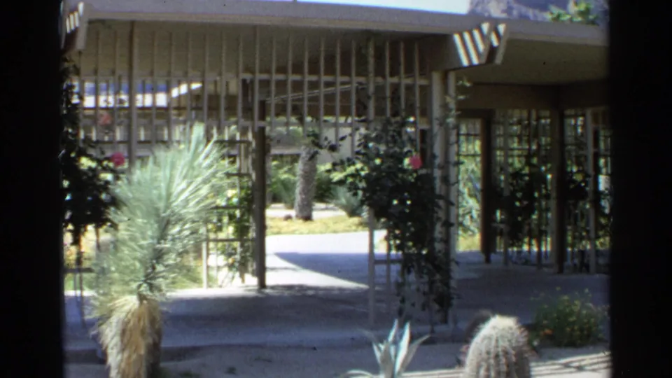 1968 Scottsdale courtyard frame with cactus, agave-like plants, screen fencing, shaded facade, and a doorway at right.