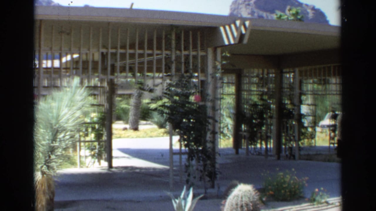 Covered modernist entrance with slatted beams, glass walls, desert plants, and rock formation beyond.