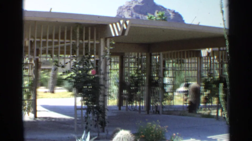 1968 Scottsdale scene showing a shaded entrance, vertical slats, flowering vines, desert plants, and a distant visitor near the building.