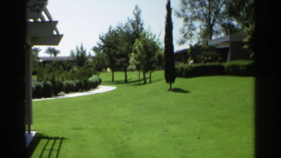 Scottsdale 1968 frame of a lush lawn bordered by trees, shrubs, a curved walkway, and part of a low building.
