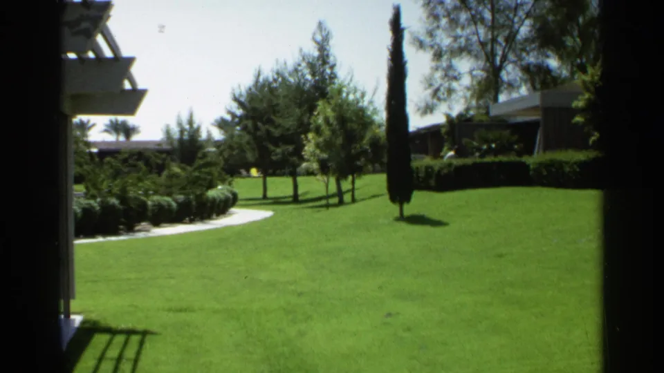 1968 Scottsdale home-movie frame showing a manicured green lawn, tall narrow tree, hedges, and low modern buildings.