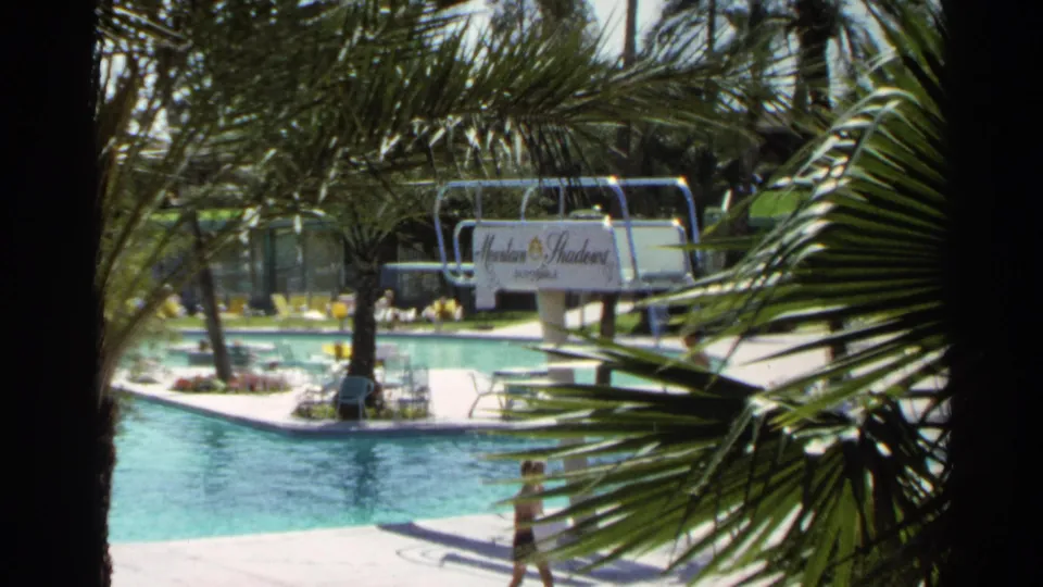 1968 Scottsdale home-movie frame showing a wider resort pool view with palm fronds, Mountain Shadows sign, seating, and a person crossing the deck.