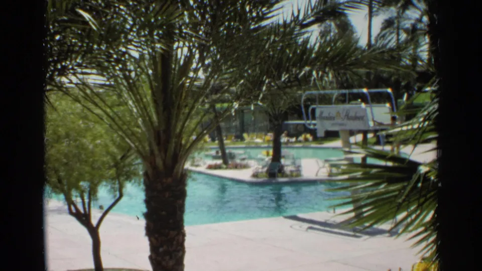 1968 home-movie view of a Scottsdale resort pool, with a palm trunk foreground, turquoise water, lounge chairs, and a distant diving board.