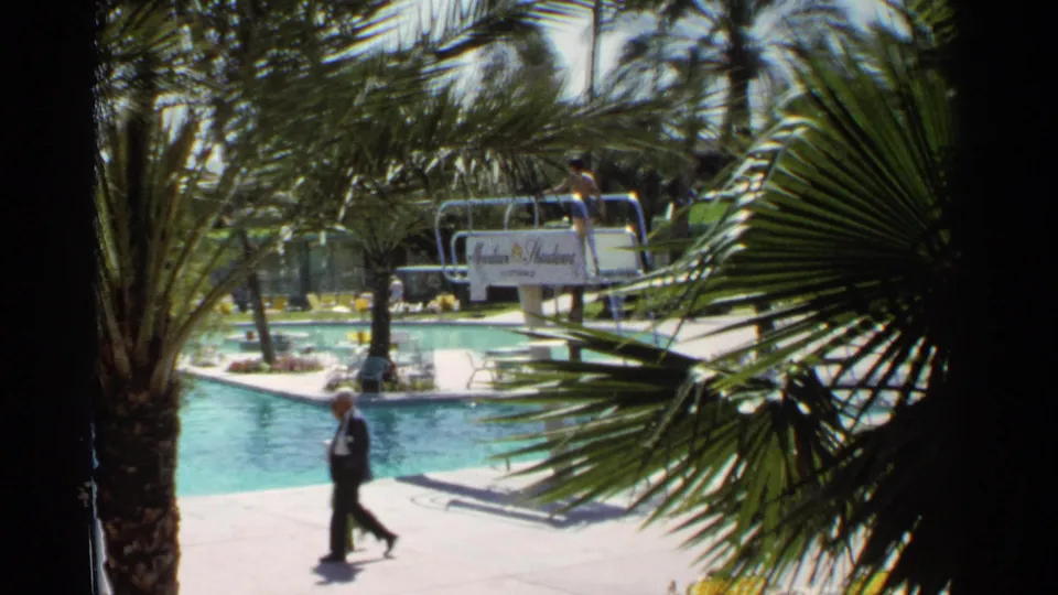1968 Scottsdale poolside frame showing an adult walking near the resort pool, partly screened by palm fronds and a visible sign area.