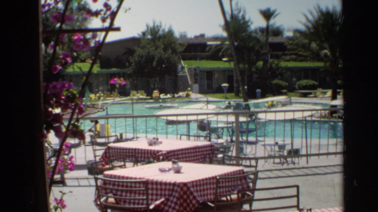 Poolside patio with checkered tables, railings, flowers, lounge chairs, and low buildings beyond the pool.