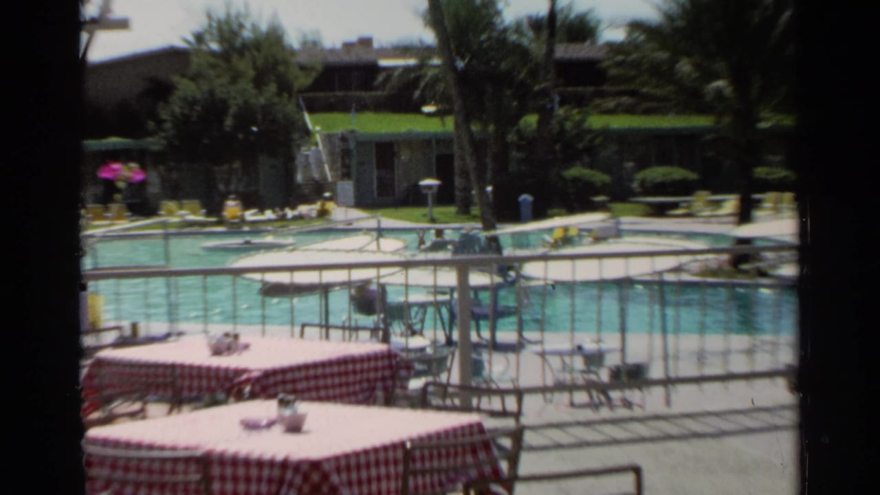 Pool area with checkered tablecloths, palm trees, railings, and lounge chairs.