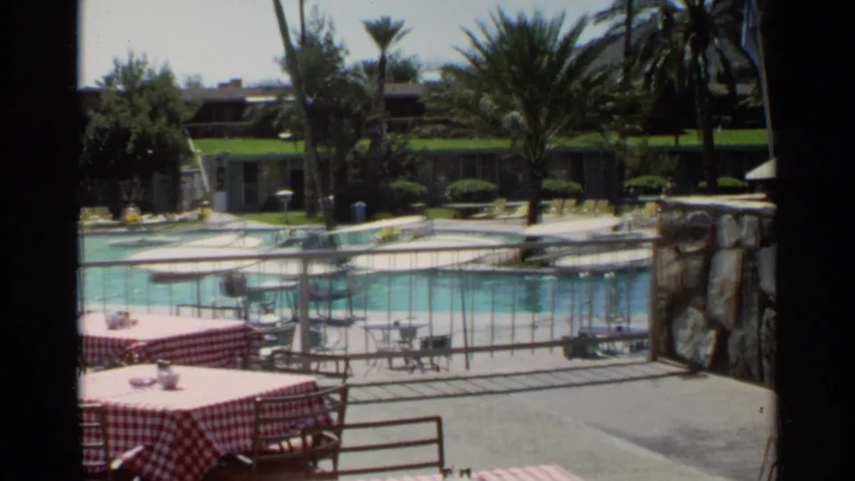 1968 Scottsdale patio view with red checkered tables, turquoise pool water, metal fencing, palms, stone wall, and low buildings beyond.
