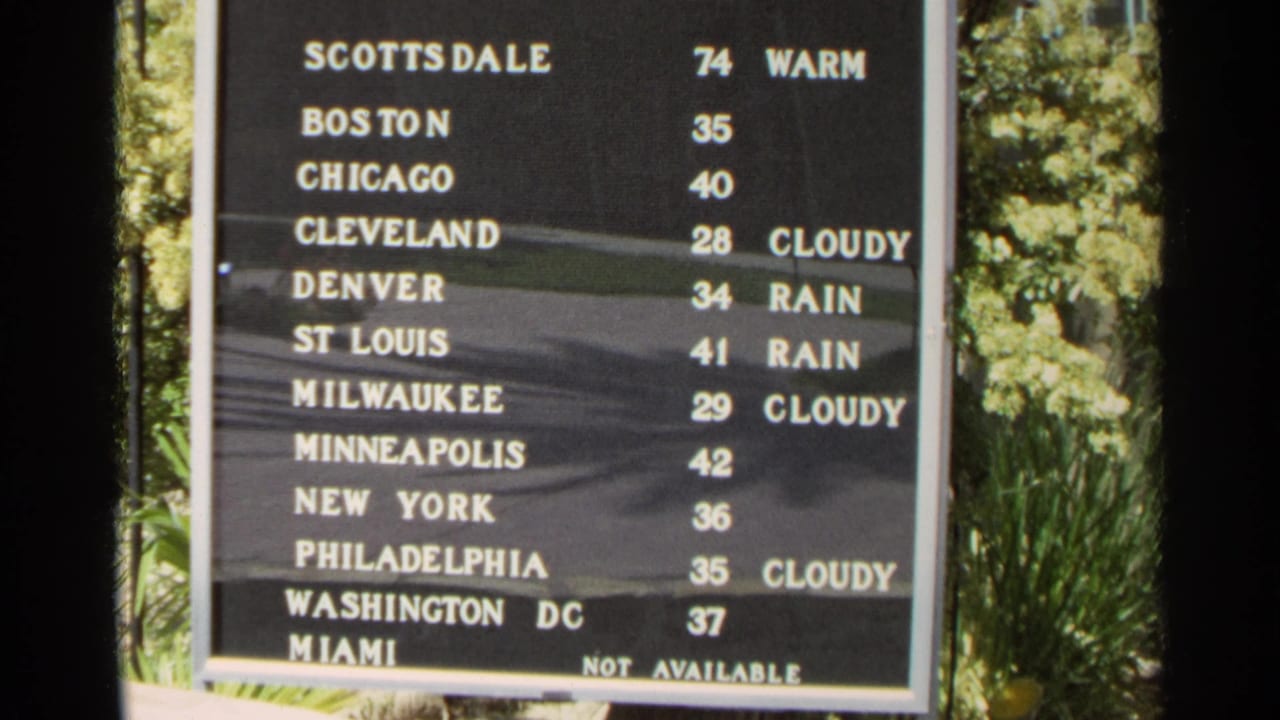 Black weather board listing U.S. cities and temperatures beside green foliage.