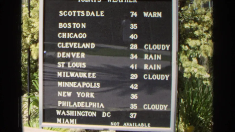Outdoor 1968 Scottsdale home-movie frame with a black lettered weather board and greenery along the side.