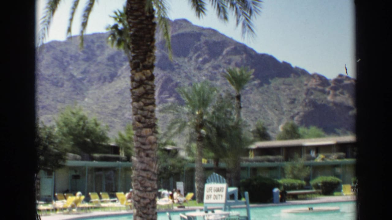 Resort pool with palm trees, lounge chairs, motel building, mountains, and a lifeguard off-duty sign.