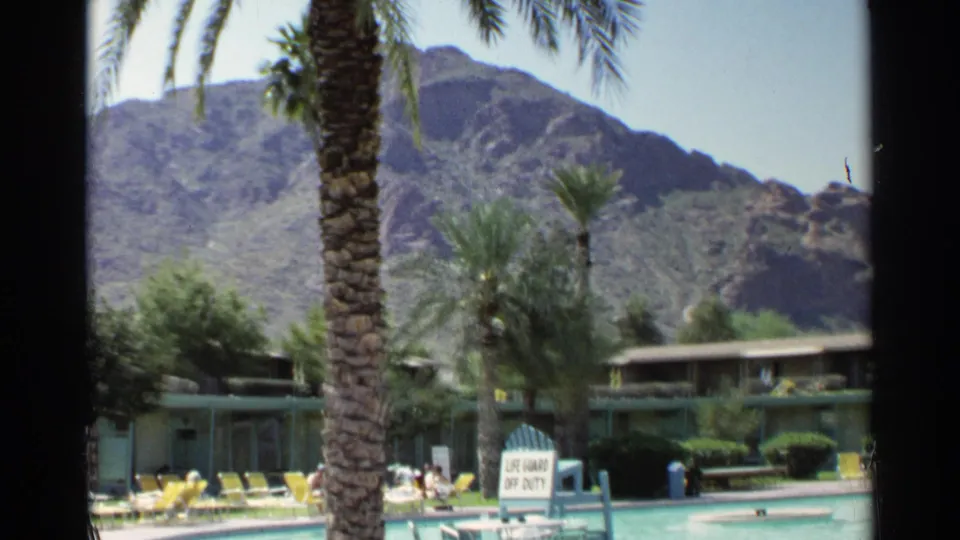 1968 Scottsdale resort pool scene with palms, yellow lounge chairs, low motel rooms, desert mountains, and a lifeguard off-duty sign.