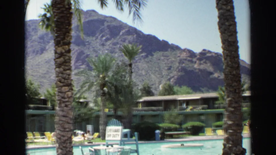 1968 Scottsdale poolside resort view showing guests near lounge chairs, palm trees, low motel rooms, and rugged desert mountains.