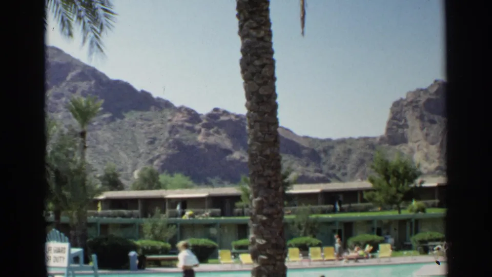 1968 Scottsdale resort pool with a person walking near the deck, guests seated nearby, palms, yellow chairs, and mountains behind.