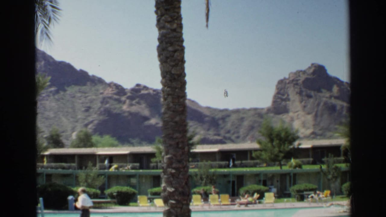 1968 Scottsdale resort pool scene with palm trunks in front, yellow loungers near turquoise water, and rocky desert mountains beyond.