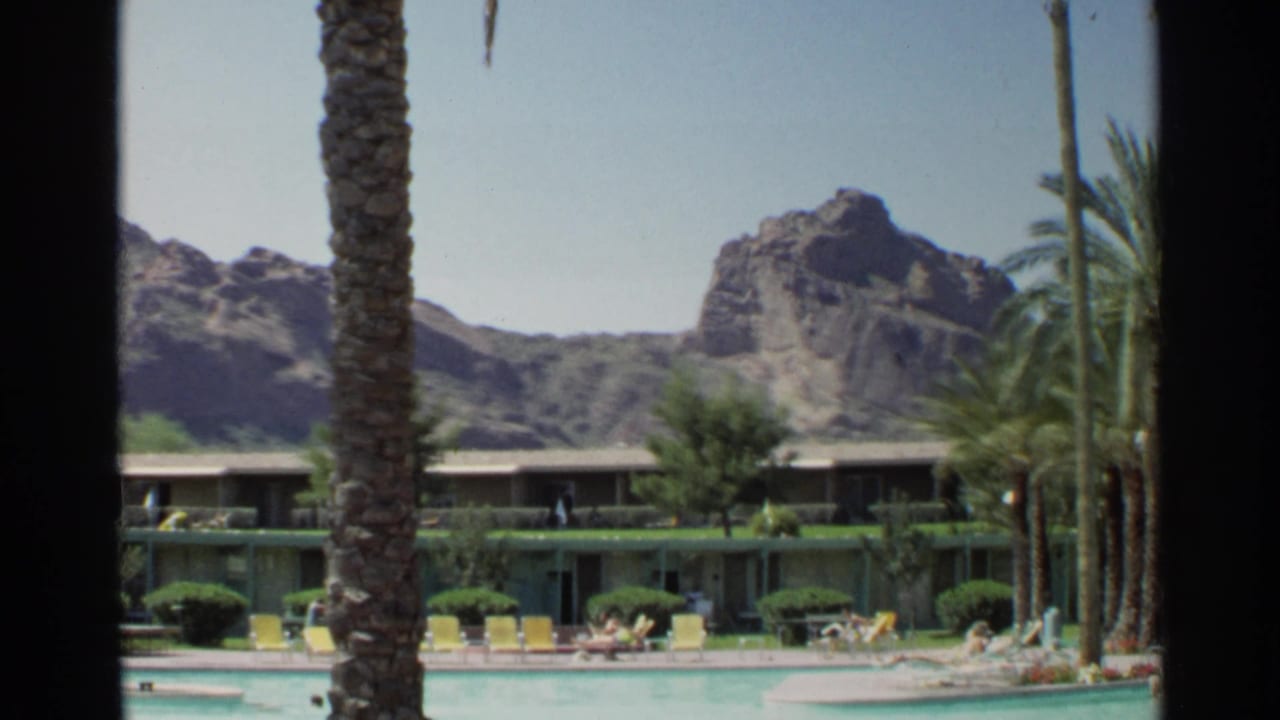 1968 Scottsdale poolside view showing a palm trunk, low resort rooms, yellow chairs, distant guests, and desert mountains.