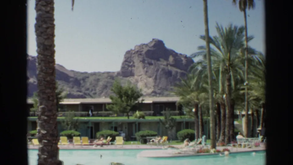 1968 Scottsdale resort pool patio with palms, yellow loungers, trimmed greenery, low rooms, and rugged mountains behind.