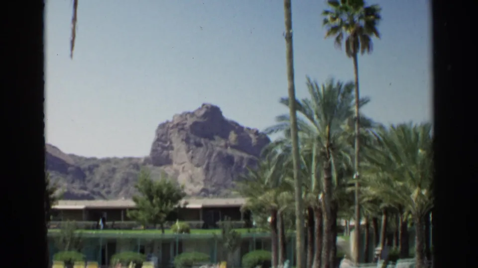 1968 Scottsdale resort pool view with clustered palms, yellow loungers, a low building, and sunlit desert mountains in back.