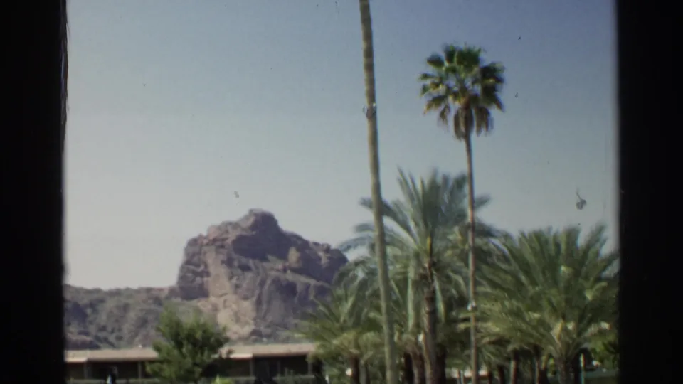 Scottsdale home-movie still with a rocky butte, palms, a low mid-century roofline, and visible film grain.