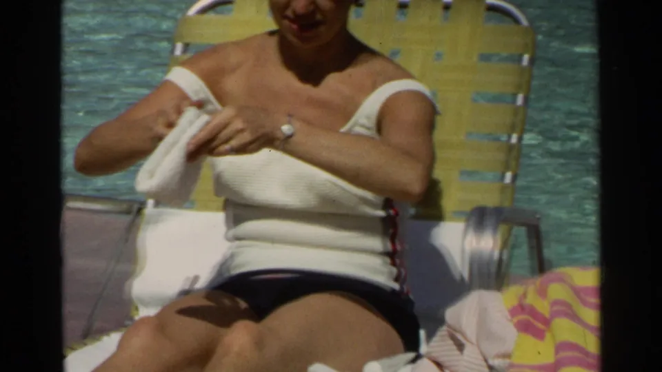 Poolside 1968 Scottsdale home-movie frame showing an adult woman seated with a towel, yellow lounge chair, and resort pool water behind her.
