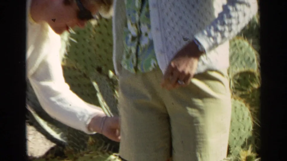The frame shows two women beside prickly pear cactus, one reaching down as the other stands in a cardigan and patterned blouse.