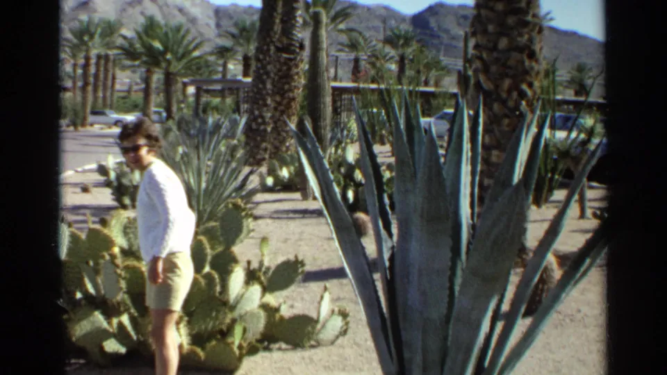 1968 Scottsdale home-movie frame of a woman in a white top and light shorts walking through a desert garden with cactus, palms, and mountains.