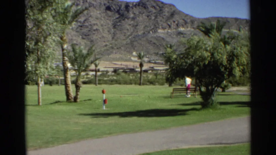 1968 Scottsdale golf-course grounds show palms, open grass, a paved path, rugged desert mountains, and a distant person in a light top and pink pants.