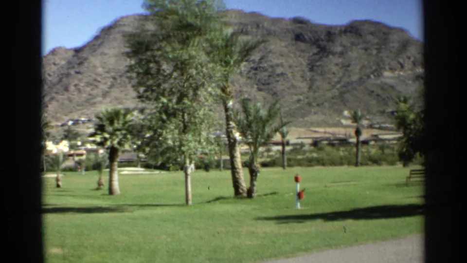 1968 Scottsdale 8mm frame showing a manicured lawn, palm trees, distant low buildings, and a rocky mountain seen through dark framing.