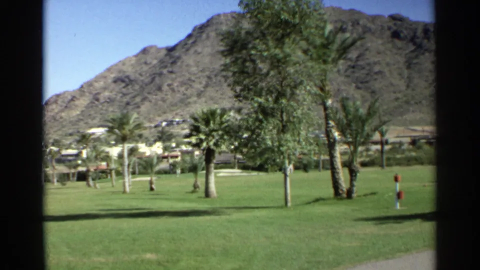 1968 Scottsdale park view with green grass, palms, a bench, low buildings, and a rugged mountain under a clear sky.