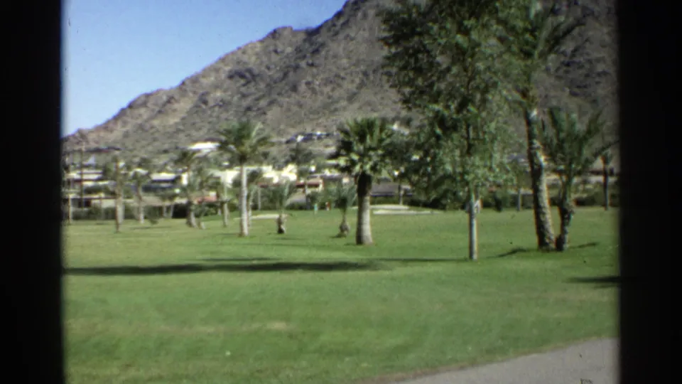 Scottsdale 1968 home-movie frame of a manicured green lawn with palms, low buildings, and desert mountain terrain beyond.