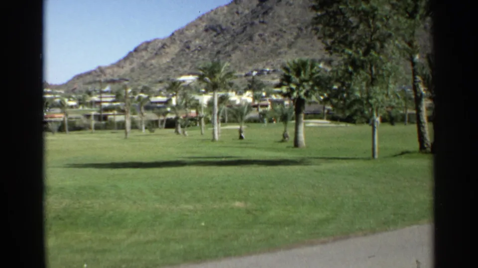 1968 Scottsdale exterior view across a bright green lawn with palms, trees, distant buildings, and a rocky mountain backdrop.
