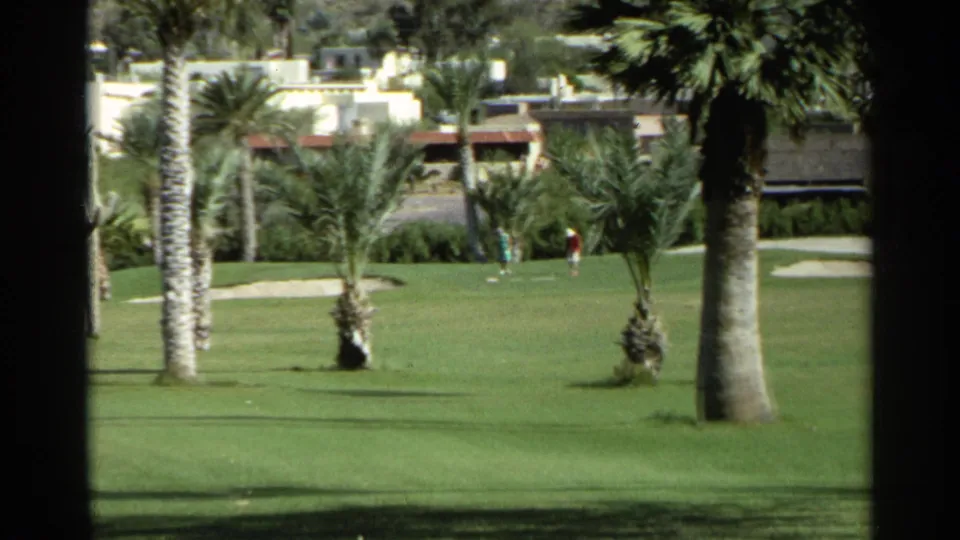 Wide 1968 Scottsdale fairway view showing palm trees, desert plants, distant golfers, buildings, and mountain backdrop.