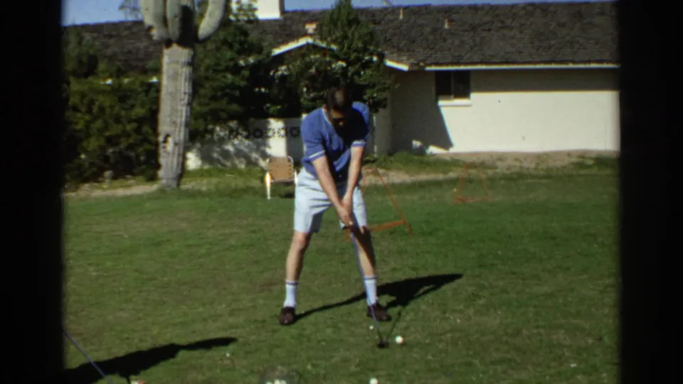 Golfer in 1968 Scottsdale footage prepares a shot on grass, with a palm trunk, lawn equipment, and low building behind him.