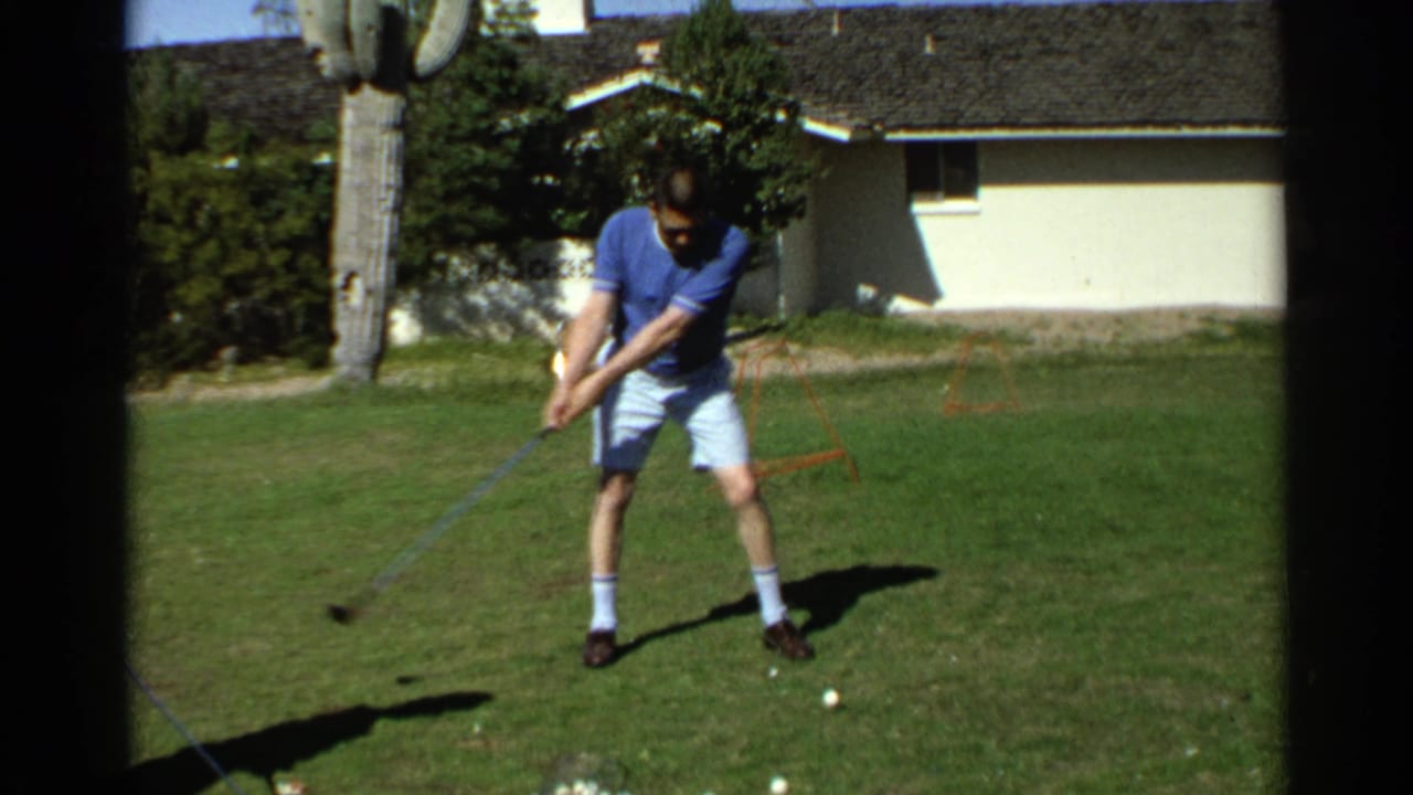 Man in sunglasses addresses a golf ball on a sunny lawn near a house and saguaro cactus.