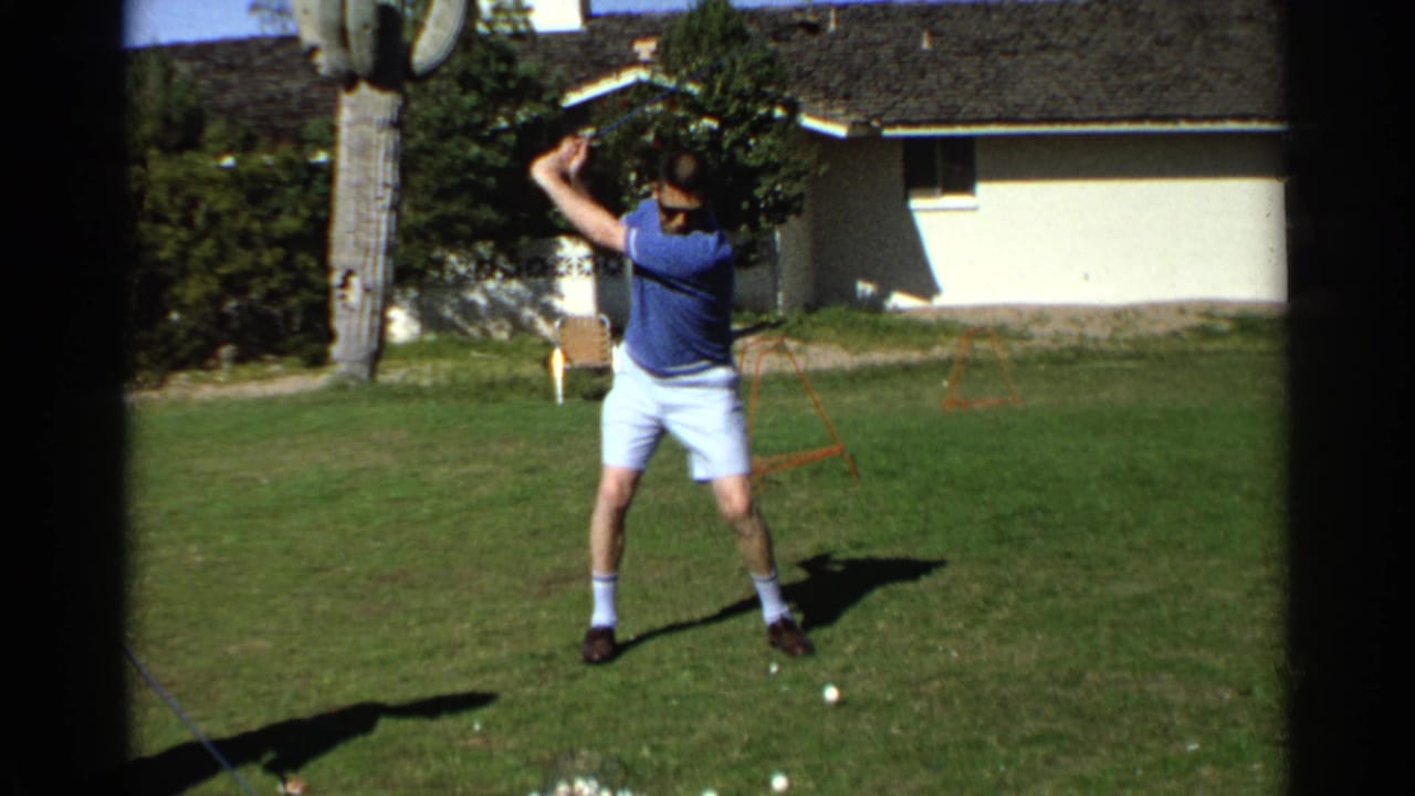 Man raises a golf club during his backswing on a lawn beside a house and cactus.