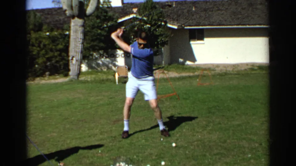 In 1968 Scottsdale, the man lifts his golf club into a backswing while practicing on a sunny residential lawn.