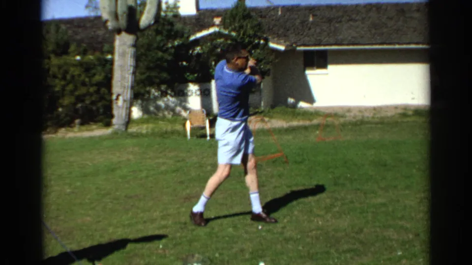 In 1968 Scottsdale, the man completes a golf swing follow-through in a sunny yard near a single-story house.