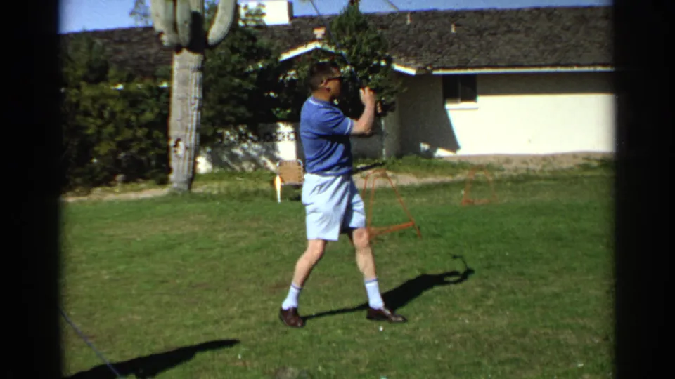 In 1968 Scottsdale, the man holds the finish of a backyard golf swing beside visible golf balls and a saguaro.