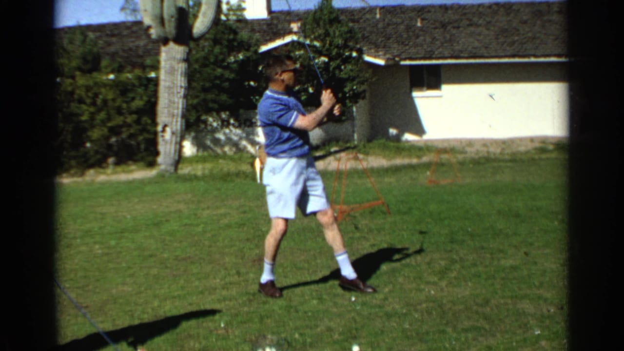 Adult man in a blue shirt swings a golf club on a backyard lawn near a saguaro cactus.