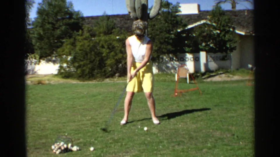 In 1968 Scottsdale, an adult woman in a white sleeveless top and yellow shorts prepares to hit a golf ball in the backyard.