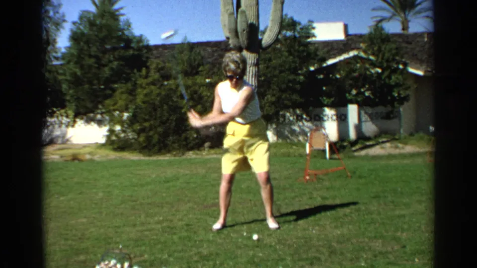 The woman swings through on a Scottsdale backyard lawn, with a saguaro cactus, golf balls, shrubs, a red chair, and a low house in view.