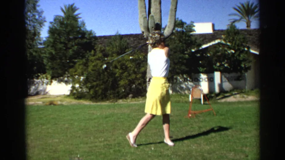 Seen from behind, the woman completes her golf swing on a green lawn in Scottsdale, with a cactus trunk, shrubs, chair, and house wall nearby.