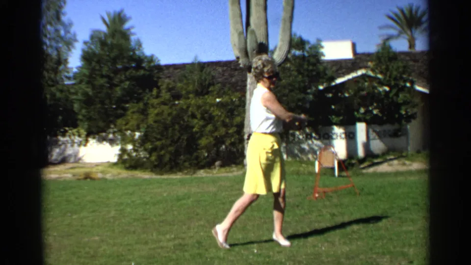 At the end of the swing, the woman steps across the Scottsdale backyard lawn with the golf club extended and a cactus, chair, shrubs, and house behind her.