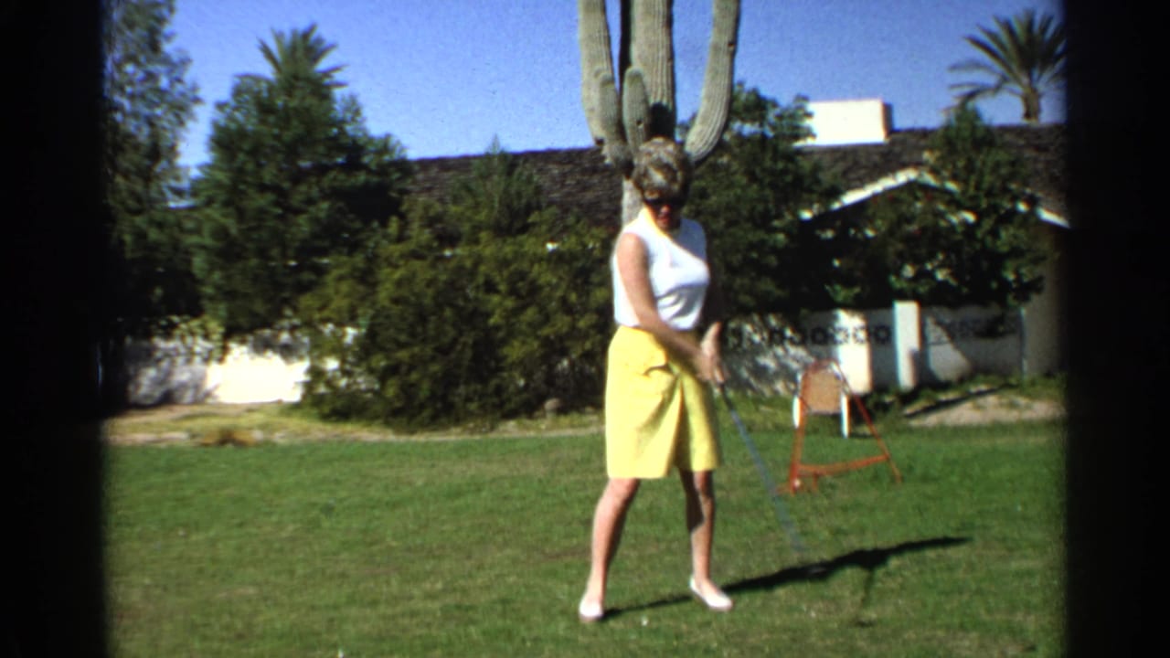 In Scottsdale, Arizona, 1968, a woman in a white sleeveless top and yellow skirt stands with a golf club on a lawn near a saguaro cactus.